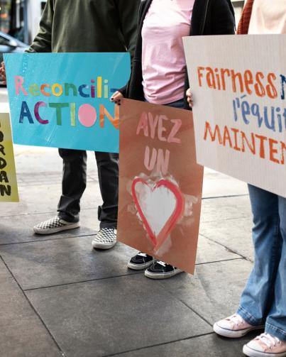 what you can do photo of children standing outside holding colourful hand painted signs reading, fairness now, l’équité maintenance, ayes un coeur, reconciliation and Jordan